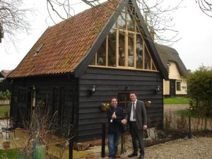 Two men stand shaking hands outside a small, dark wooden house with a steep red-tiled roof and large windows. Another house and trees are visible in the background on a cloudy day.