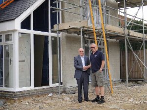 Two men are shaking hands in front of a house under construction, with scaffolding and building materials visible around them. One man is formally dressed in a suit, while the other wears casual work clothes.