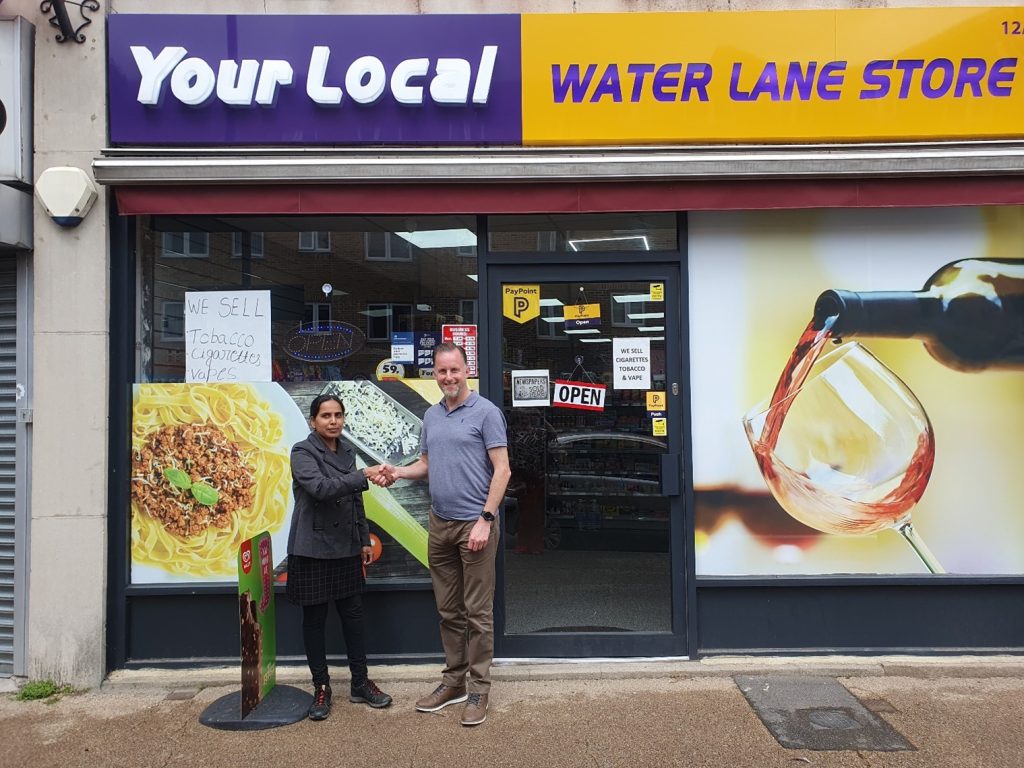 Two people are shaking hands and smiling in front of an off-licence called Your Local Water Lane Store. The shopfront displays images of pasta and wine and has several signs in the windows.