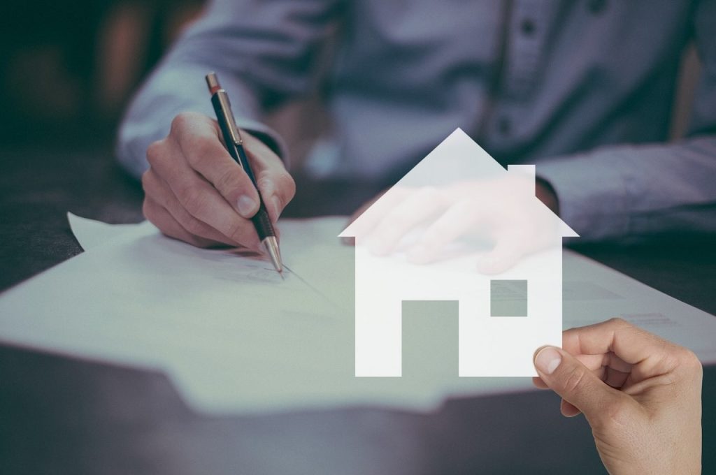 Development finance - ASC A person signs a document with a pen at a desk whilst another hand holds a transparent house icon in the foreground, symbolising property or home ownership.