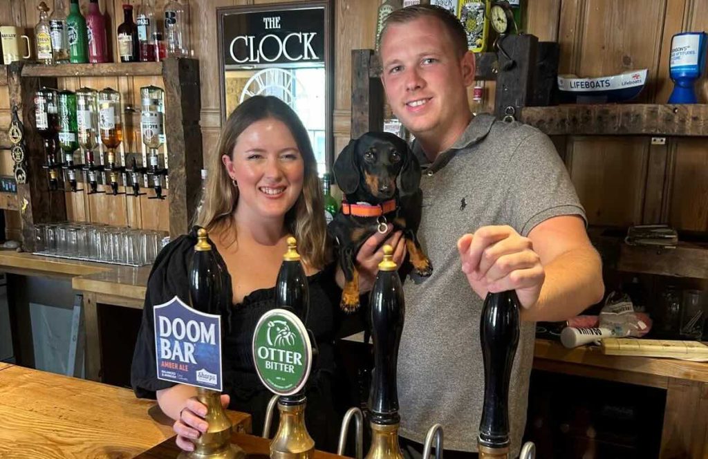 ASC brings life back to a Dorset pub - ASC A smiling woman and man stand behind a pub bar, holding a small black and brown dachshund. Ale pumps labelled Doom Bar and Otter Bitter are in front of them, with glasses and bottles on shelves behind.