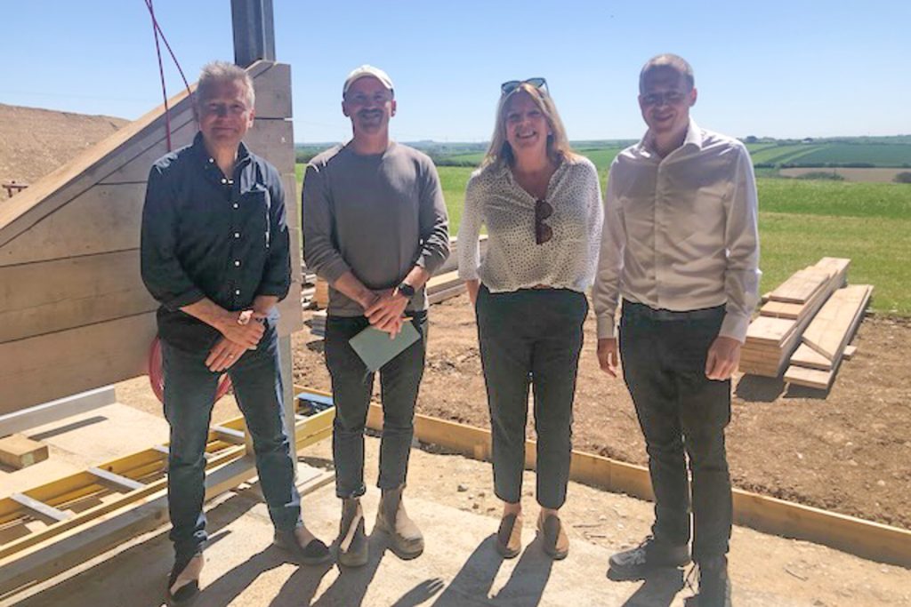 Four people stand side by side outdoors at a golf driving range, smiling at the camera. Behind them are grassy fields, building materials, and a clear blue sky.