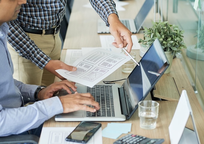 Two people work at a desk with a laptop, discussing paper sketches of a website layout. One types on the laptop while the other points at the screen with a pen. A phone, glass of water, and plant are on the desk.
