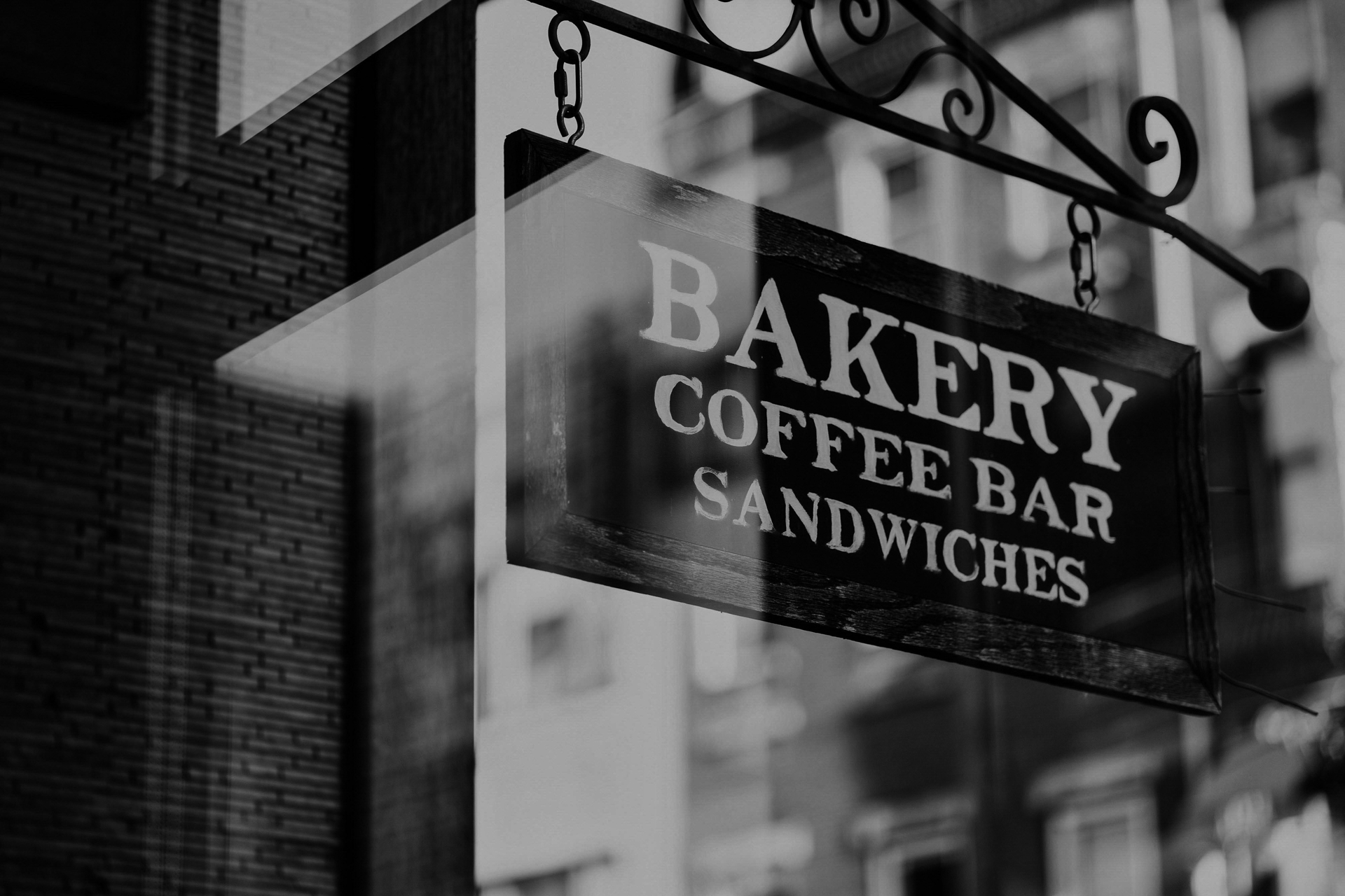 A black and white photo of a hanging sign reading “BAKERY COFFEE BAR SANDWICHES” outside a building, with blurred city buildings reflected in a nearby window.
