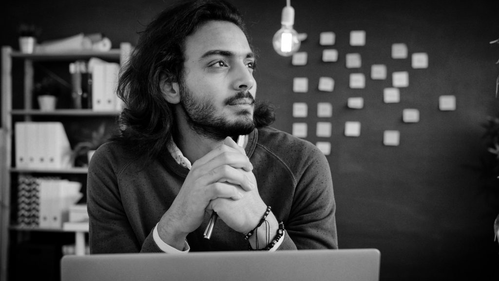 A man with long hair sits at a desk with his hands clasped, looking thoughtfully to the side. Behind him are shelves, a hanging light bulb, and a wall covered with sticky notes.