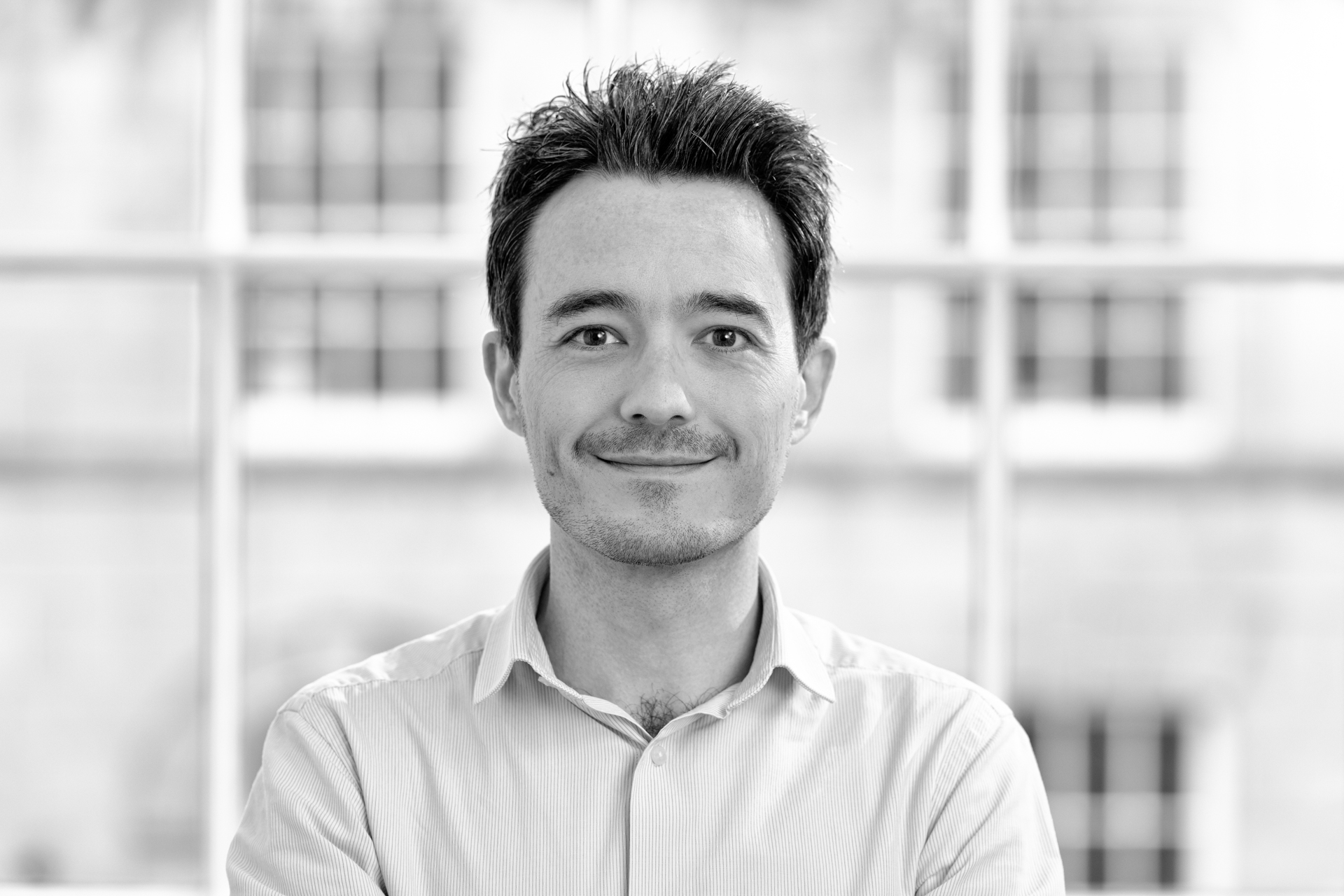 A man with short, dark hair wearing a light-coloured button-up shirt smiles at the camera. He stands indoors in front of a large window with blurred buildings in the background. The image is in black and white.