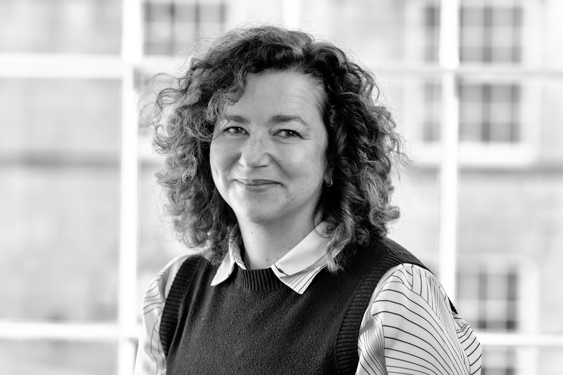 A woman with curly hair and a friendly expression is wearing a striped shirt and dark waistcoat, posing indoors in front of large windows. The photo is in black and white.