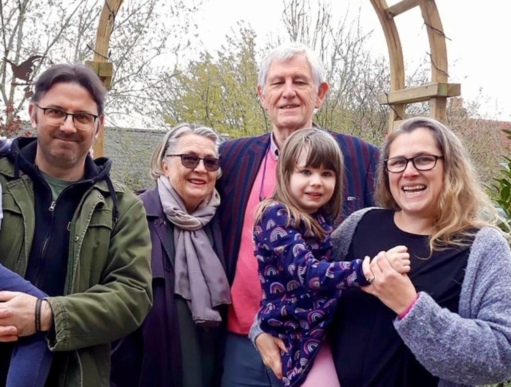 Five people, including an older couple, two adults, and a child in colourful clothes, stand closely together outdoors under a wooden arch, smiling at the camera with trees in the background.