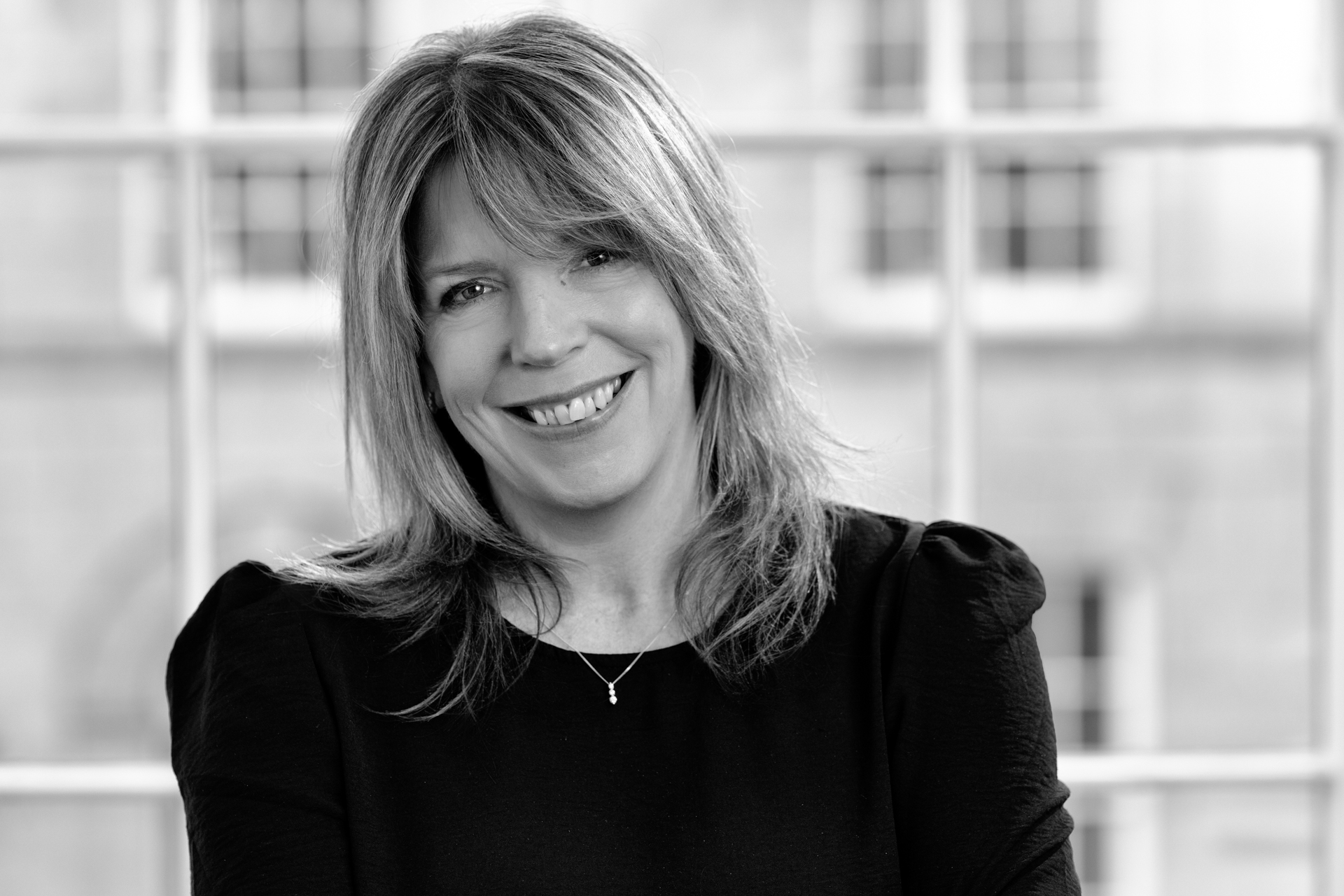 Black and white photo of a smiling woman with shoulder-length hair, wearing a dark top and a small necklace, standing in front of a window with a blurred building in the background.