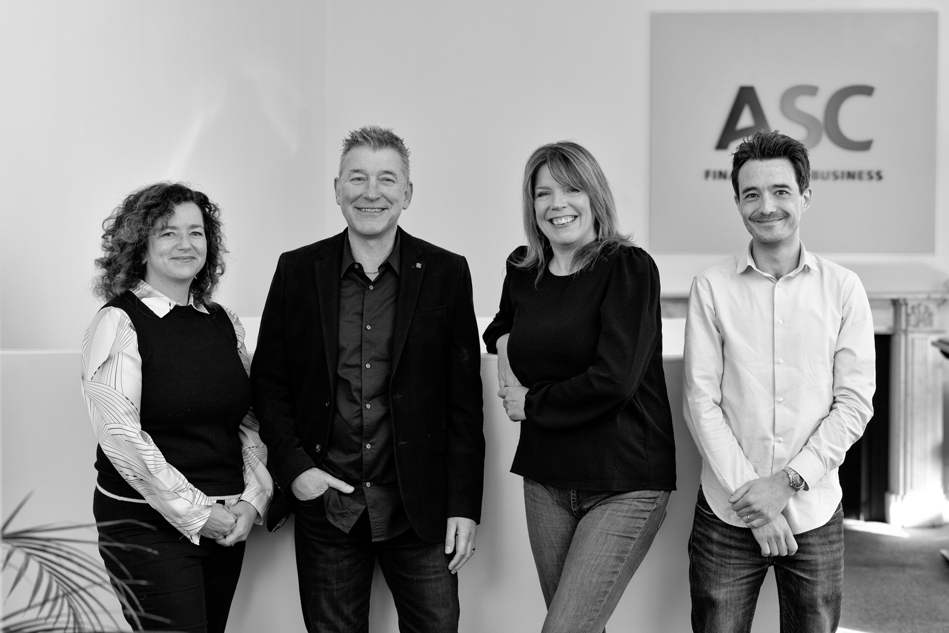 Four people stand smiling in an office setting in front of a wall with an ASC Finance for Business sign. The group includes two men and two women, casually dressed and standing close together.
