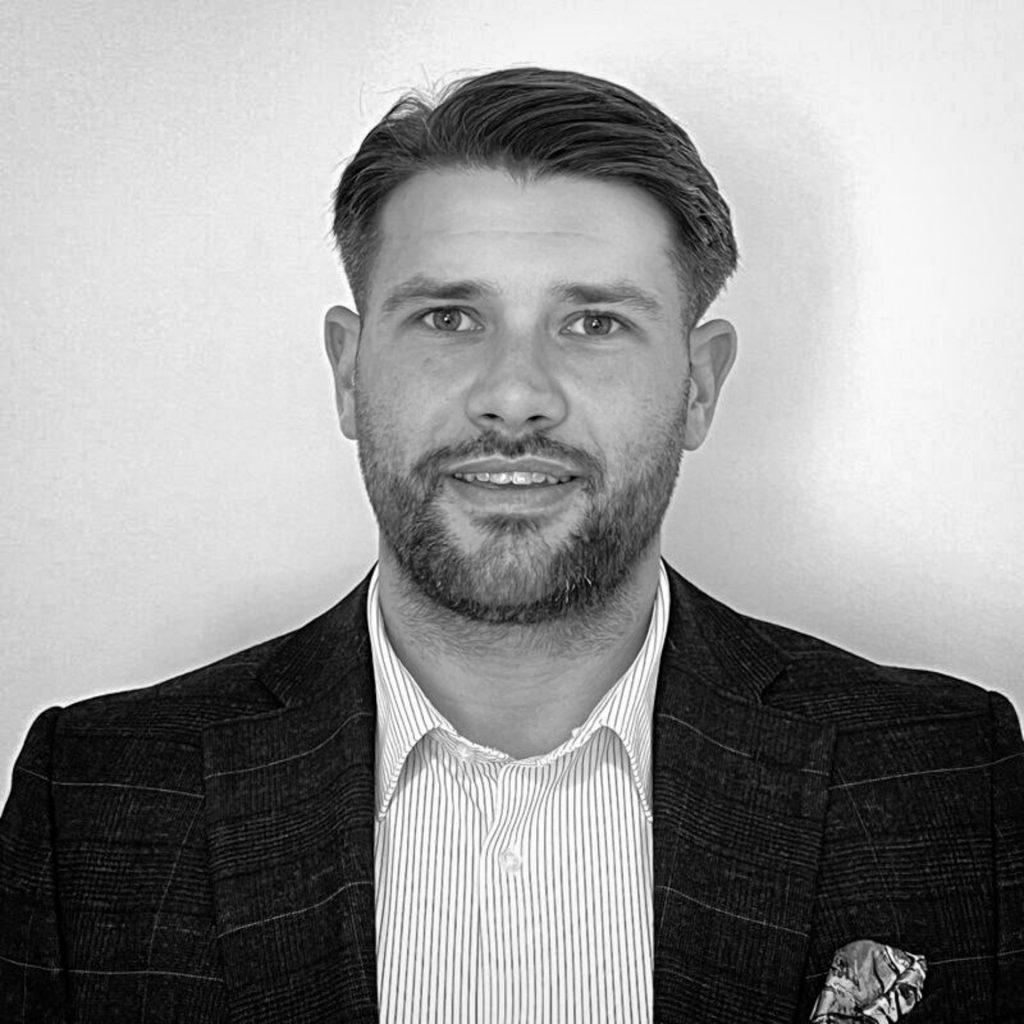 A man with short hair and a beard, wearing a striped collared shirt and a checked blazer with a pocket square, poses in front of a plain background. The image is in black and white.