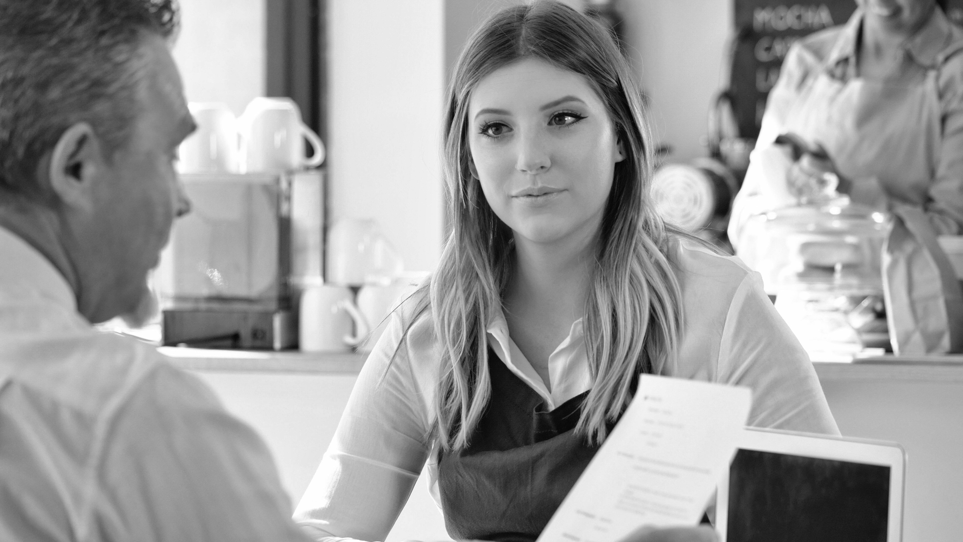 A young woman in an apron sits across a table from a man holding papers, likely in a job interview at a café. The background shows a counter, cups, and another person working.