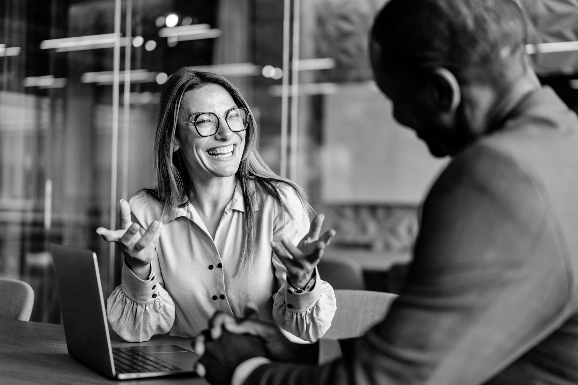 A woman wearing glasses smiles and gestures enthusiastically whilst talking to a man in an office setting. Both are seated at a table with a laptop open in front of them.