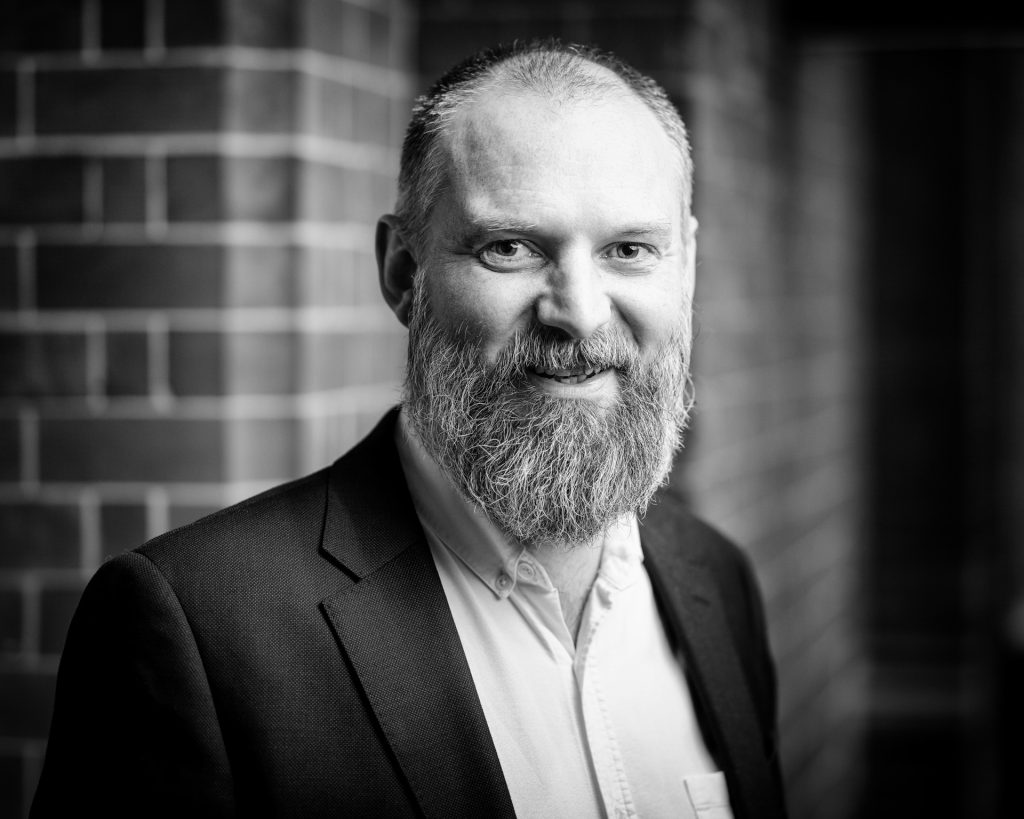 Black-and-white portrait of a bearded man in a suit jacket and collared shirt, smiling at the camera, standing in front of a brick wall backdrop.