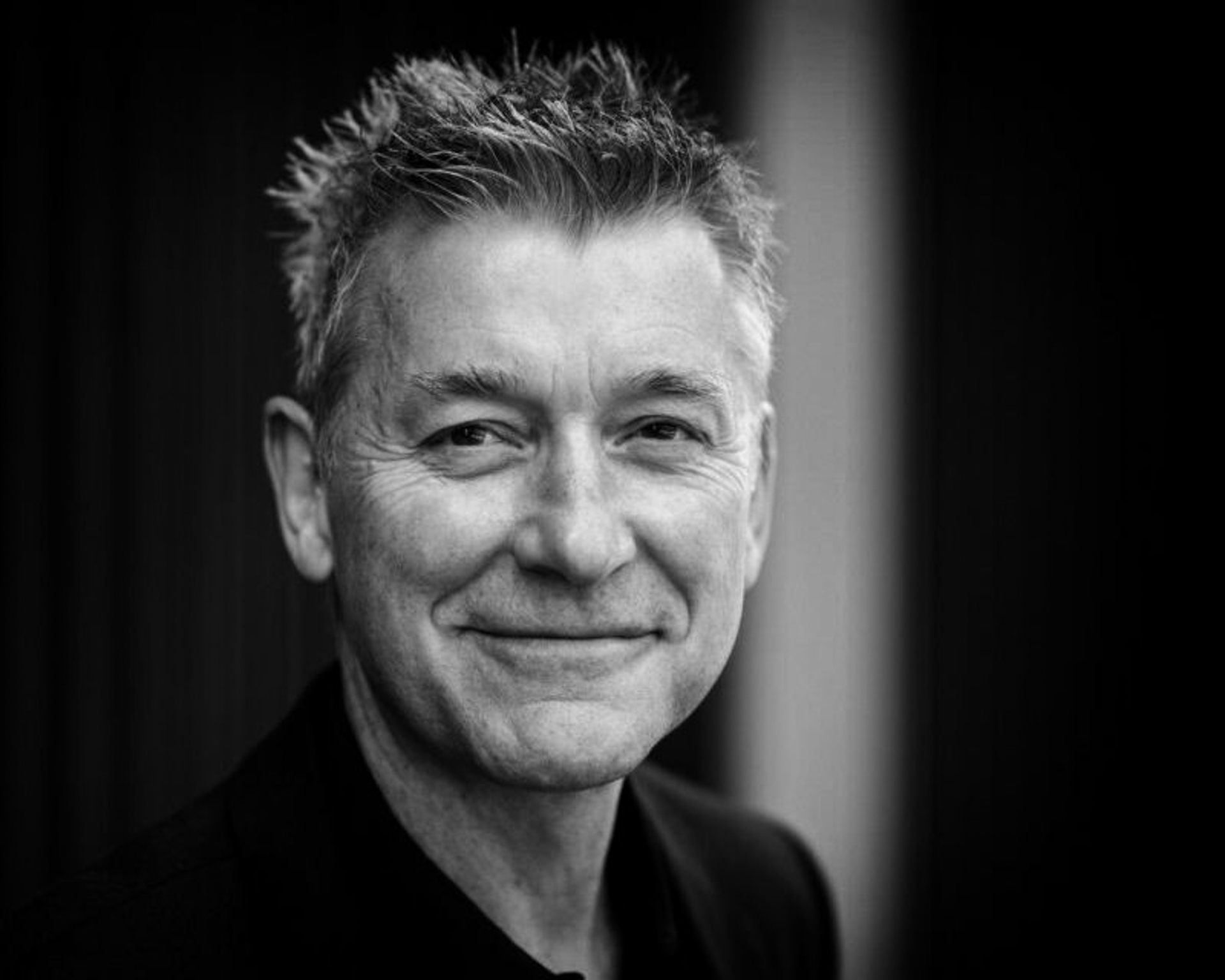 A greyscale portrait of a middle-aged man with short, spiky hair, smiling gently at the camera. He is wearing a dark shirt and the background is softly blurred.