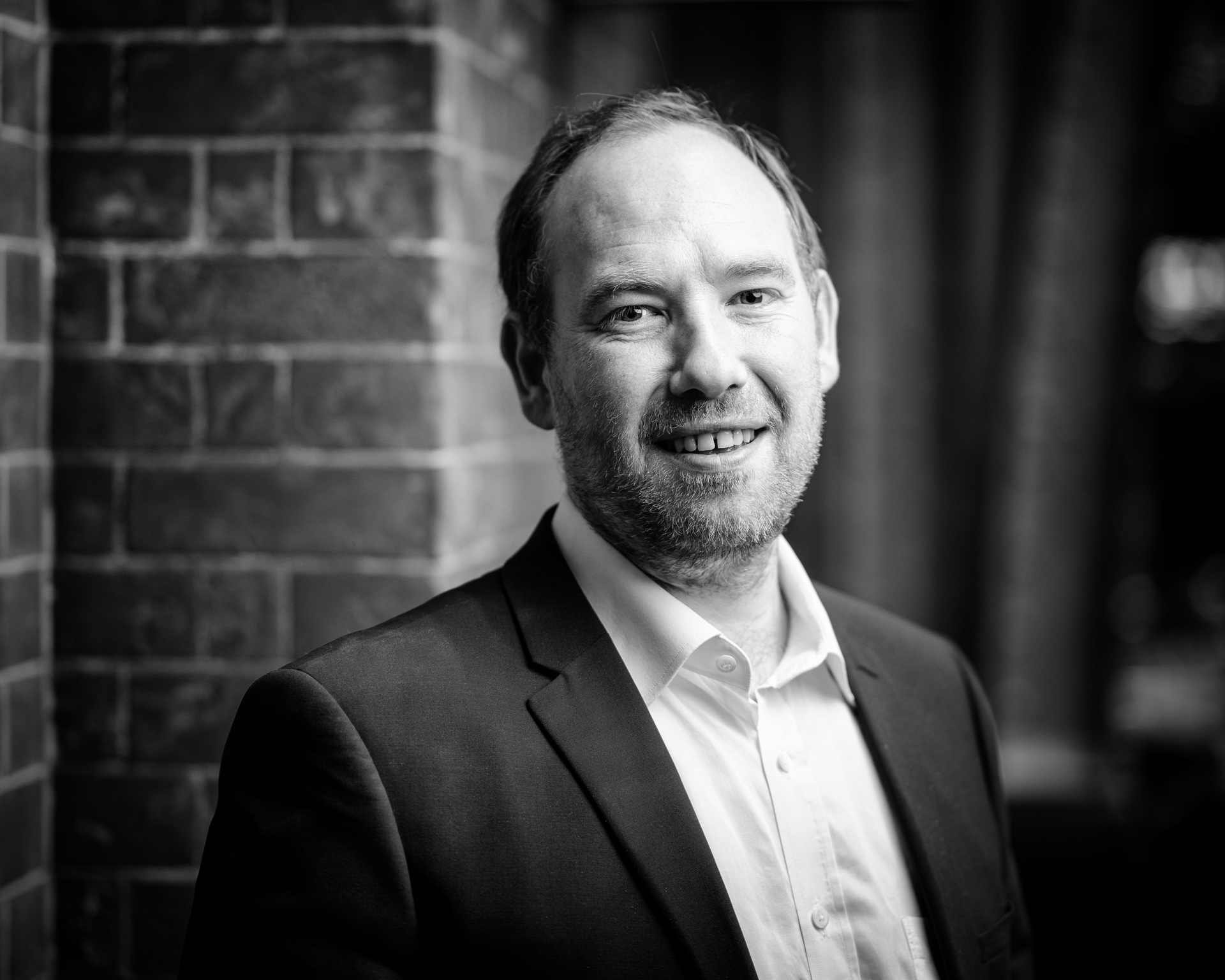 A man in a suit jacket and open-collared shirt stands in front of a brick wall, smiling. The photo is black and white with a softly blurred background.