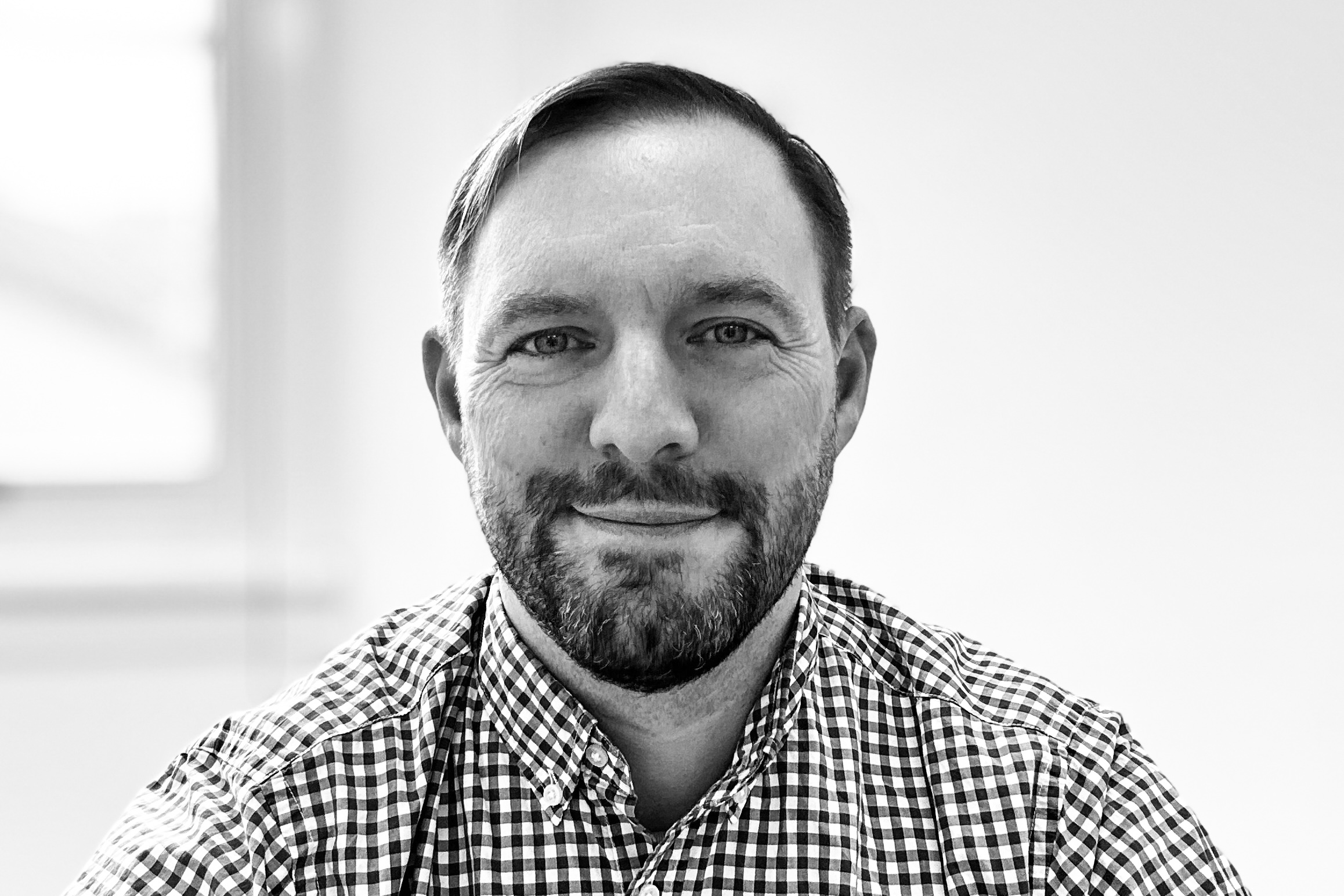 A man with short hair, a beard, and a checked shirt smiles at the camera in a black and white photo with a blurred background.