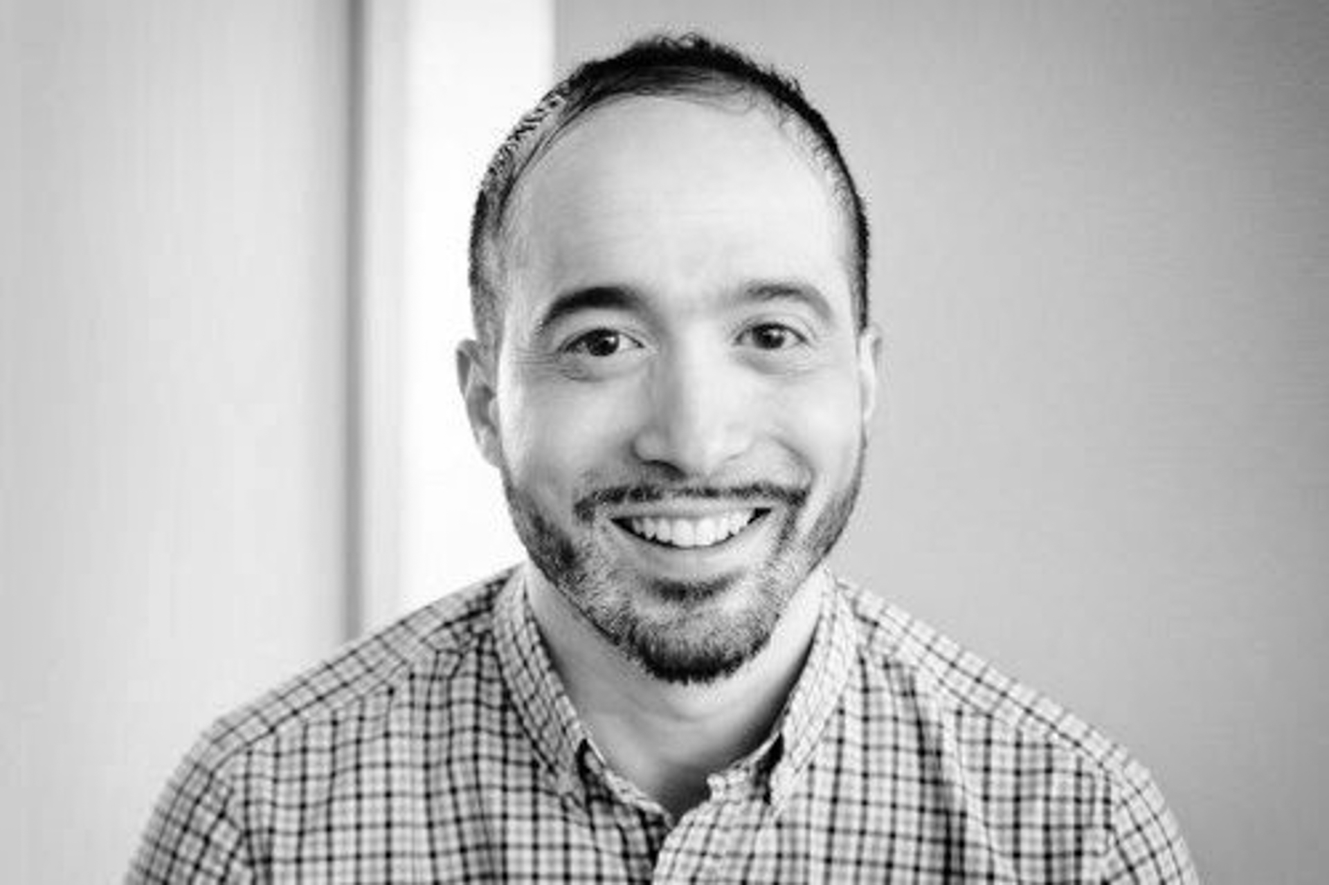 A smiling man with short hair, a beard, and a moustache, wearing a checked shirt, is looking directly at the camera. The photo is in black and white with a softly blurred background.