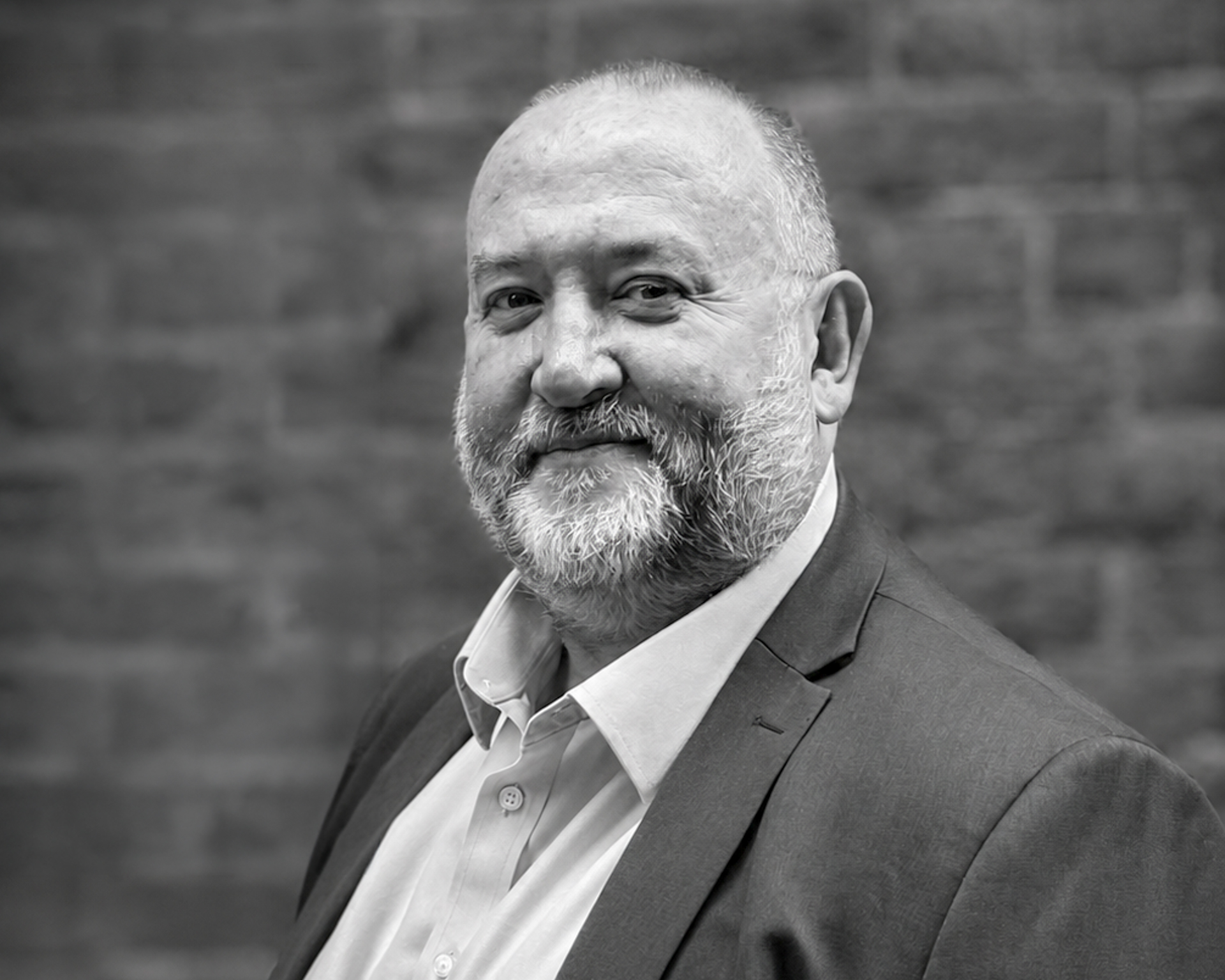 Portrait of a smiling, bearded man in a suit against a blurred brick background in grayscale.