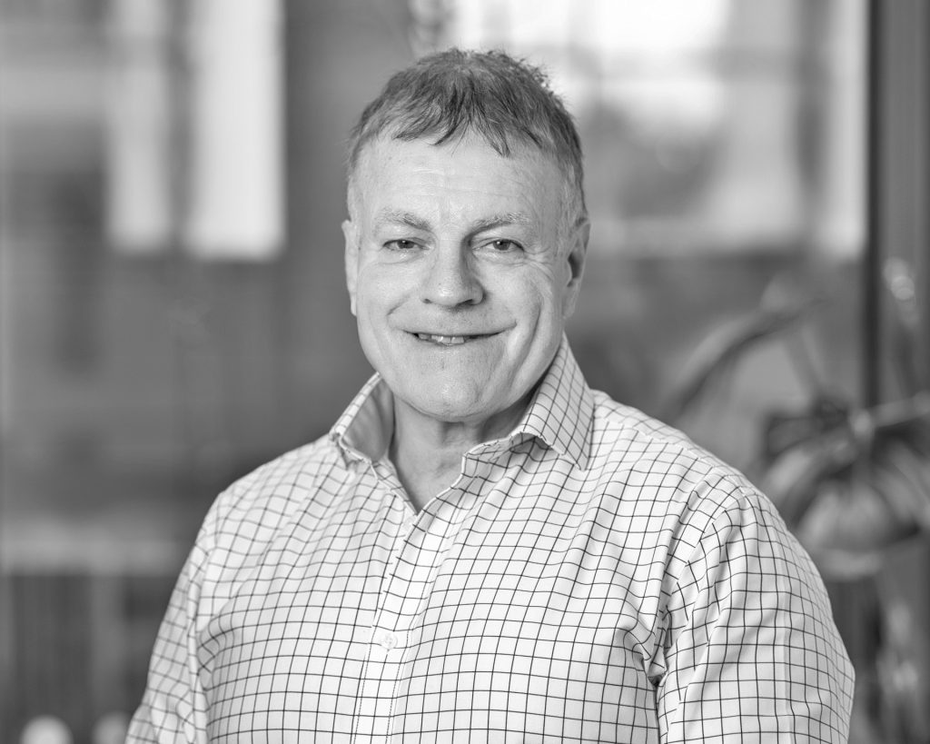A middle-aged man with short hair smiles at the camera. He is wearing a checked button-up shirt, and the background is blurred with hints of windows and indoor plants. The image is in black and white.