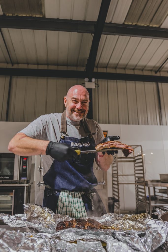 Photo of Adam , pitmaster at The Aussie Smoker, smiling at the camera and holding a tay of barbecued meat
